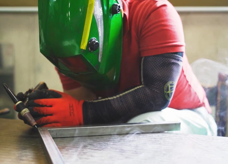 土肥板金工業の職人がTIG溶接でステンレス部材を加工している様子。溶接マスクと手袋を着用した精密作業。