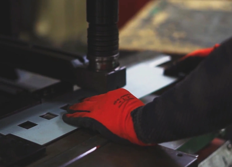 プレス機で金属板に穴あけ加工を行う職人の手元。赤い作業手袋を着用した板金加工の作業風景。
