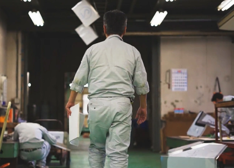 工場内で金属部品を持って歩く板金職人の後ろ姿。作業場で金物製作が進む様子。